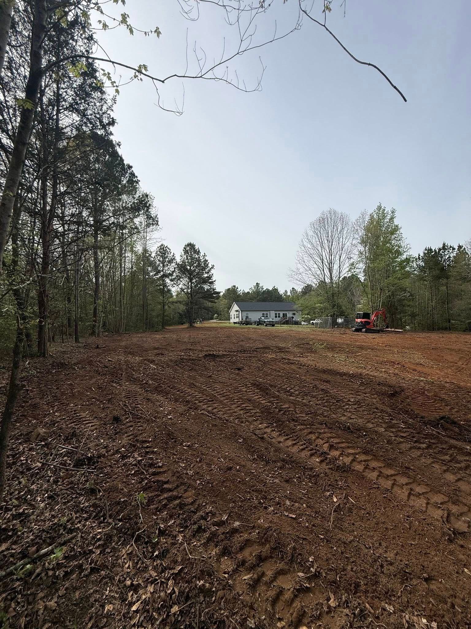 Cleared dirt lot beside trees, with a small excavator near a white building under a bright sky