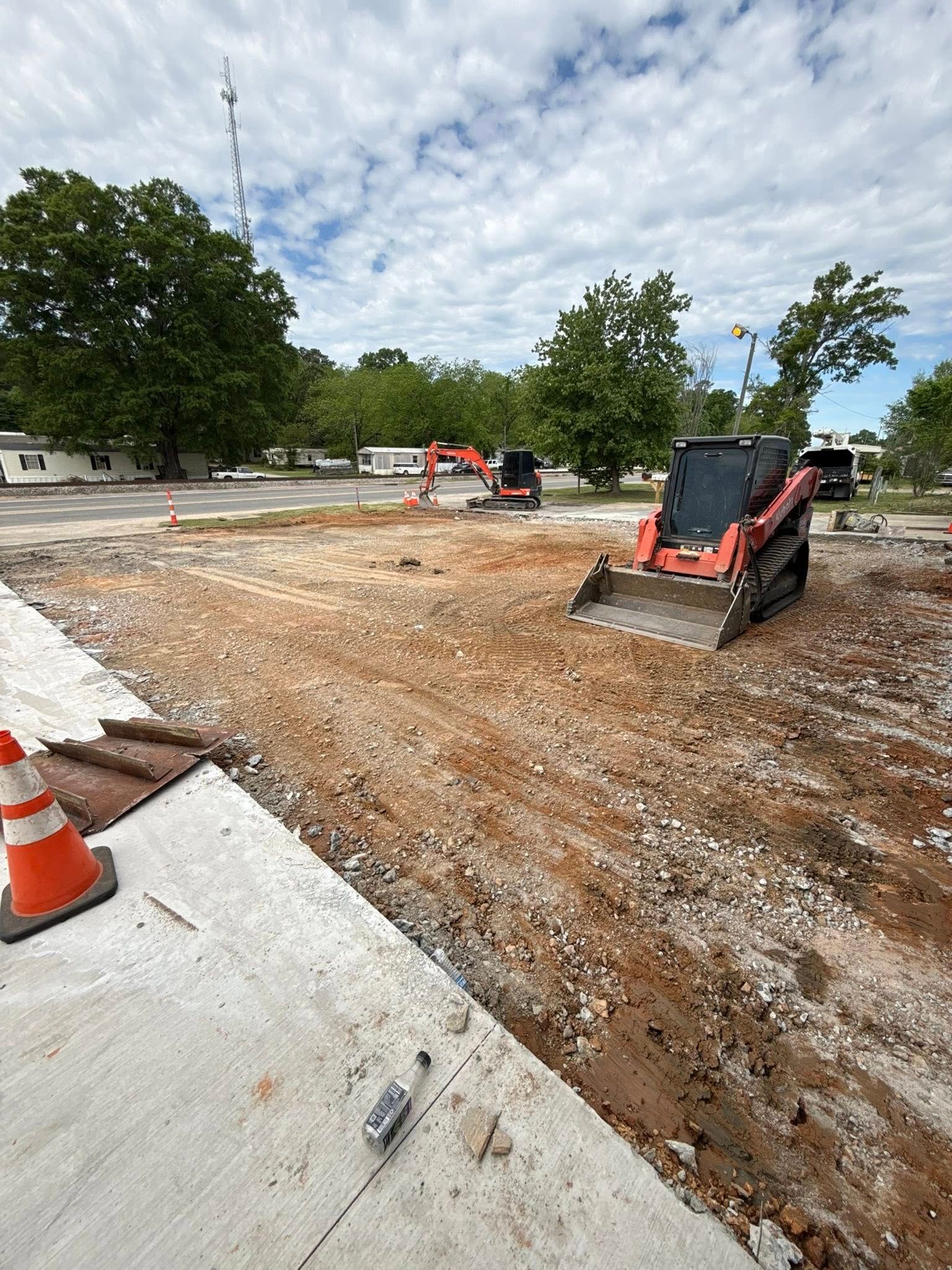 Construction site with skid steer on dirt lot, orange cones, and trees under a cloudy sky