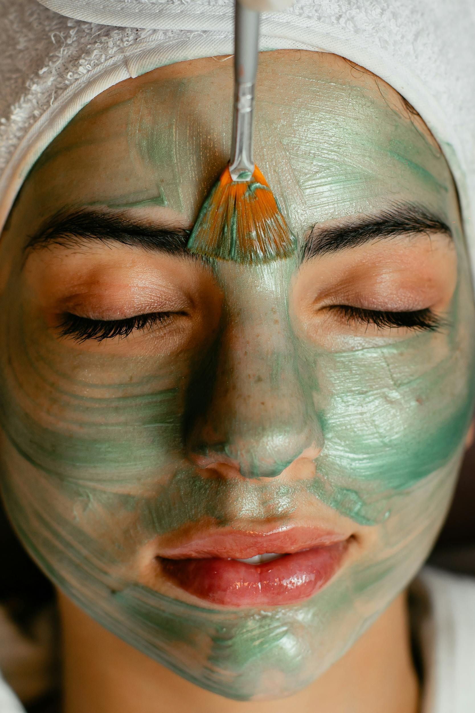 Woman with closed eyes, face covered in green mask, receiving facial treatment.