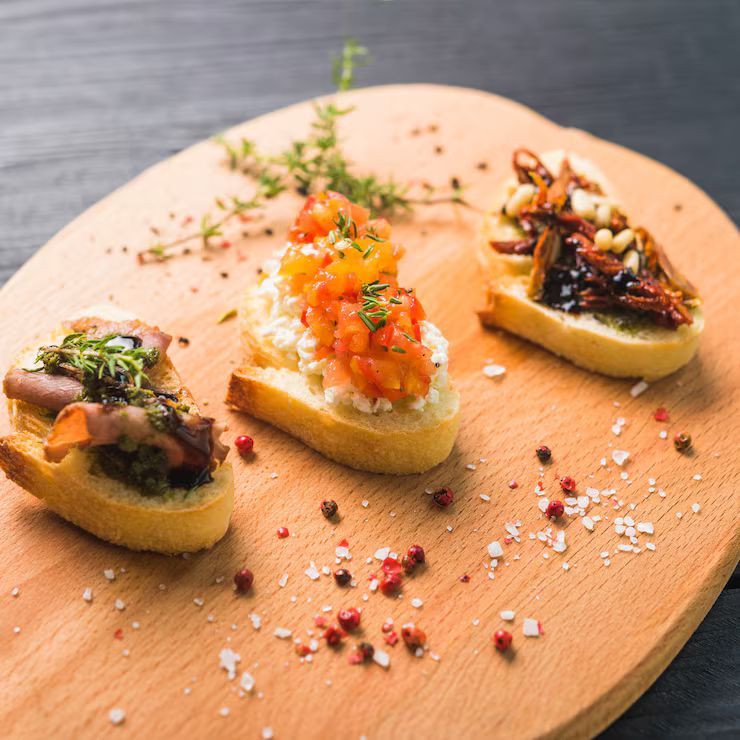 Three bruschetta appetizers on a wooden board: tomato and ricotta, pesto and cured meat, and sun-dried tomatoes.
