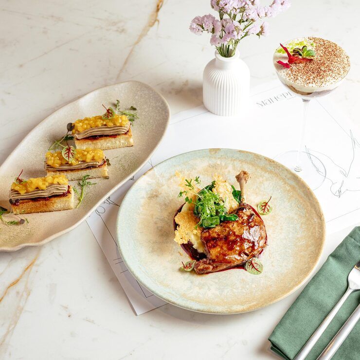Gourmet dishes on a marble table: duck leg with mashed potatoes, appetizers, dessert, and a vase of flowers.