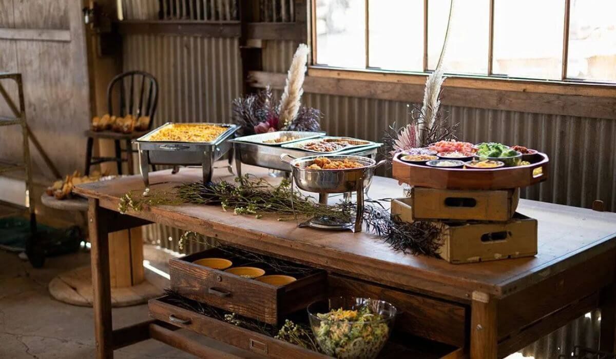 Buffet table inside a rustic barn, featuring chafing dishes with food and decorative herbs.