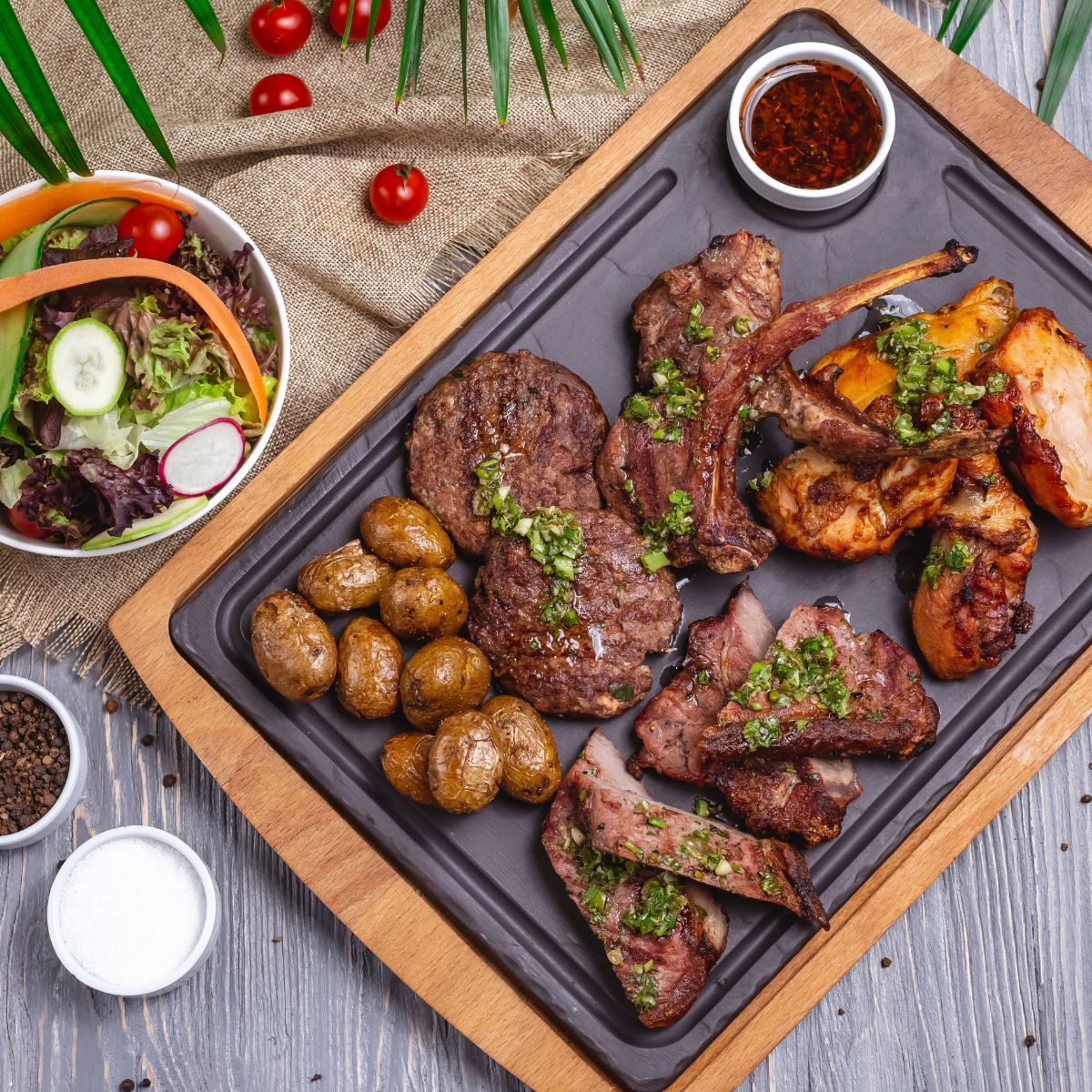 Grilled meat platter with potatoes, salad, and sauce on a wooden board.