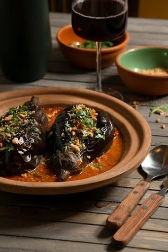 Two stuffed peppers in sauce on a rustic table with wine and utensils