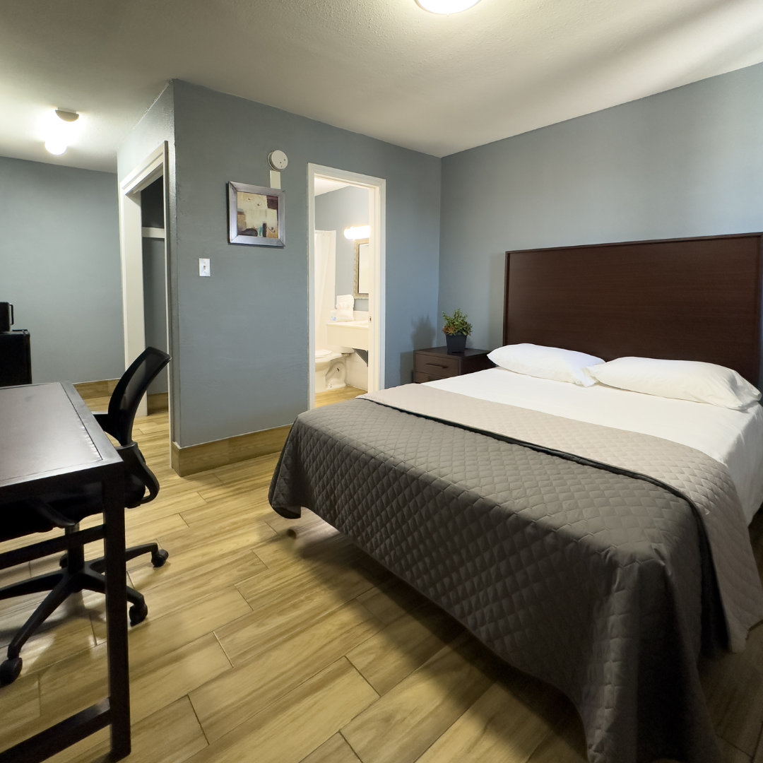Hotel room with a bed, desk, and bathroom visible in the background. Wooden floors and blue walls.