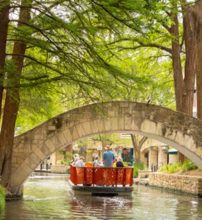 River boat with passengers passing under a stone arch bridge, surrounded by greenery.