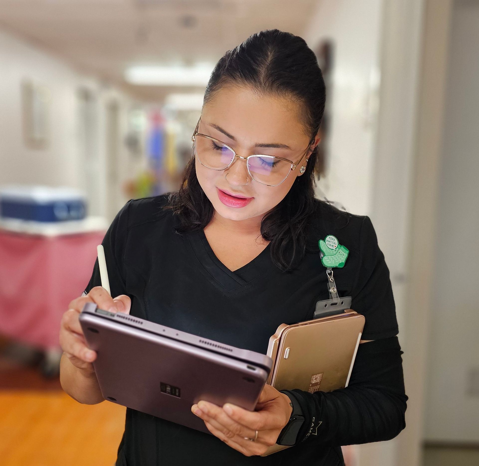A healthcare worker dressed in black scrubs stands in a clinic hallway, looking down at a tablet she's writing on with a stylus. She wears glasses and has a badge reel and a small decorative pin on her chest. She holds a second tablet or device tucked under her arm. The background shows a blurred corridor with medical equipment.