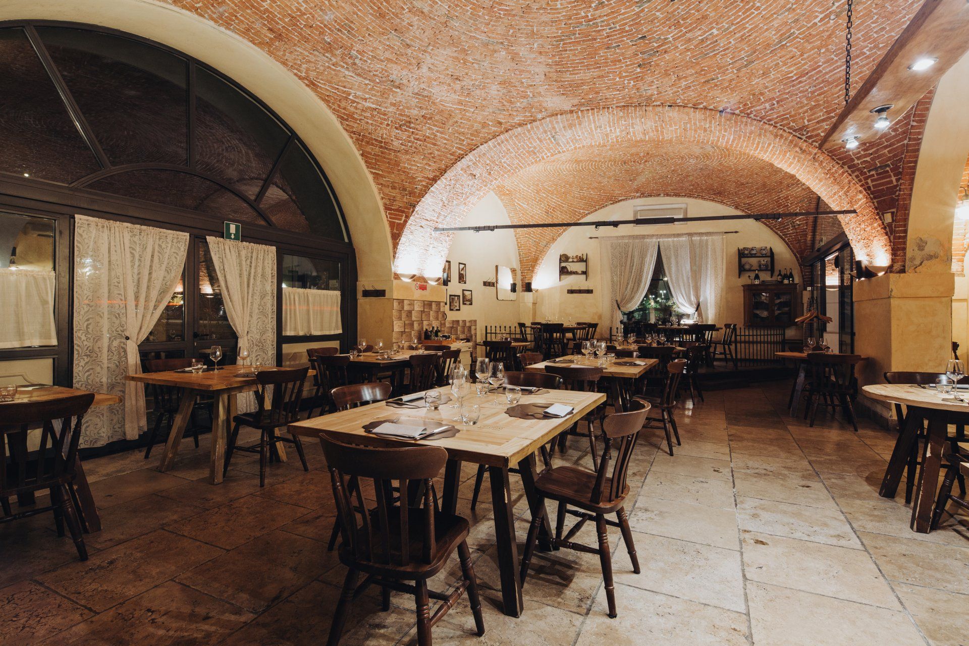 Interno del ristorante con soffitto a volta in mattoni, tavoli, sedie e finestra con tende.