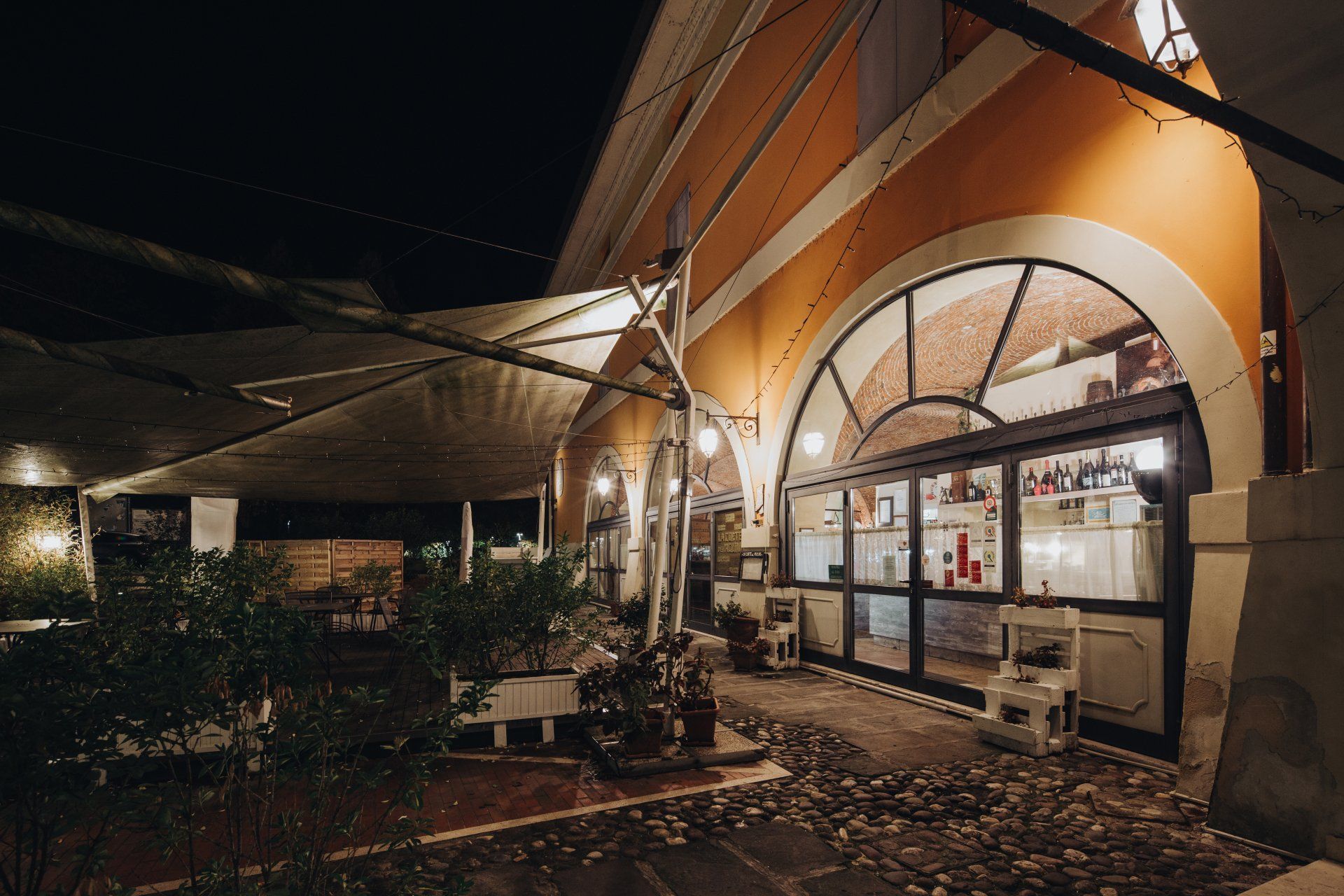 Esterno notturno di un ristorante con finestre ad arco, posti a sedere nel patio e tenda da sole.