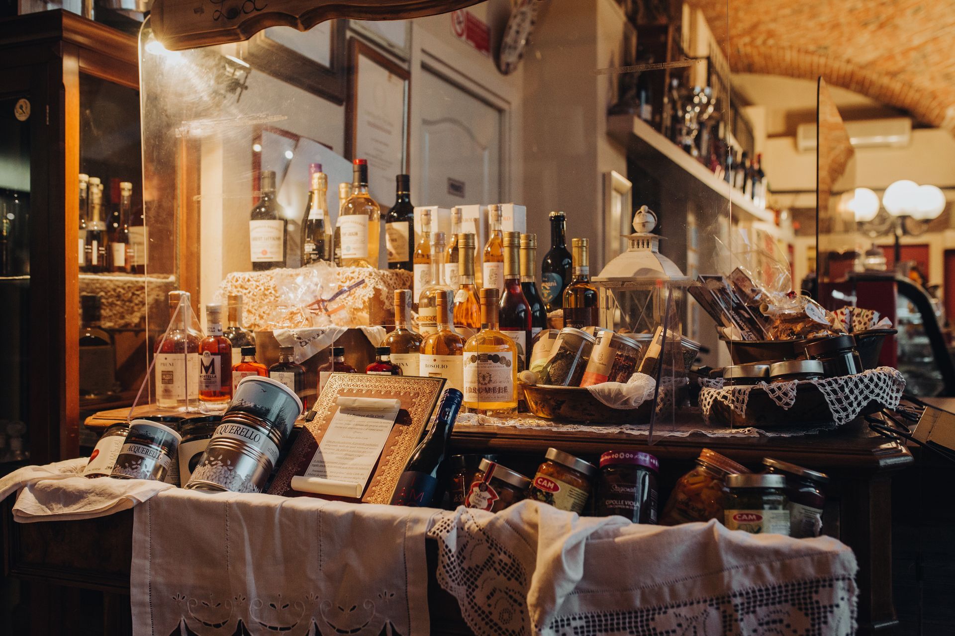 Esposizione del ristorante: bottiglie di vino e barattoli di conserve su un tavolo ricoperto di pizzo.
