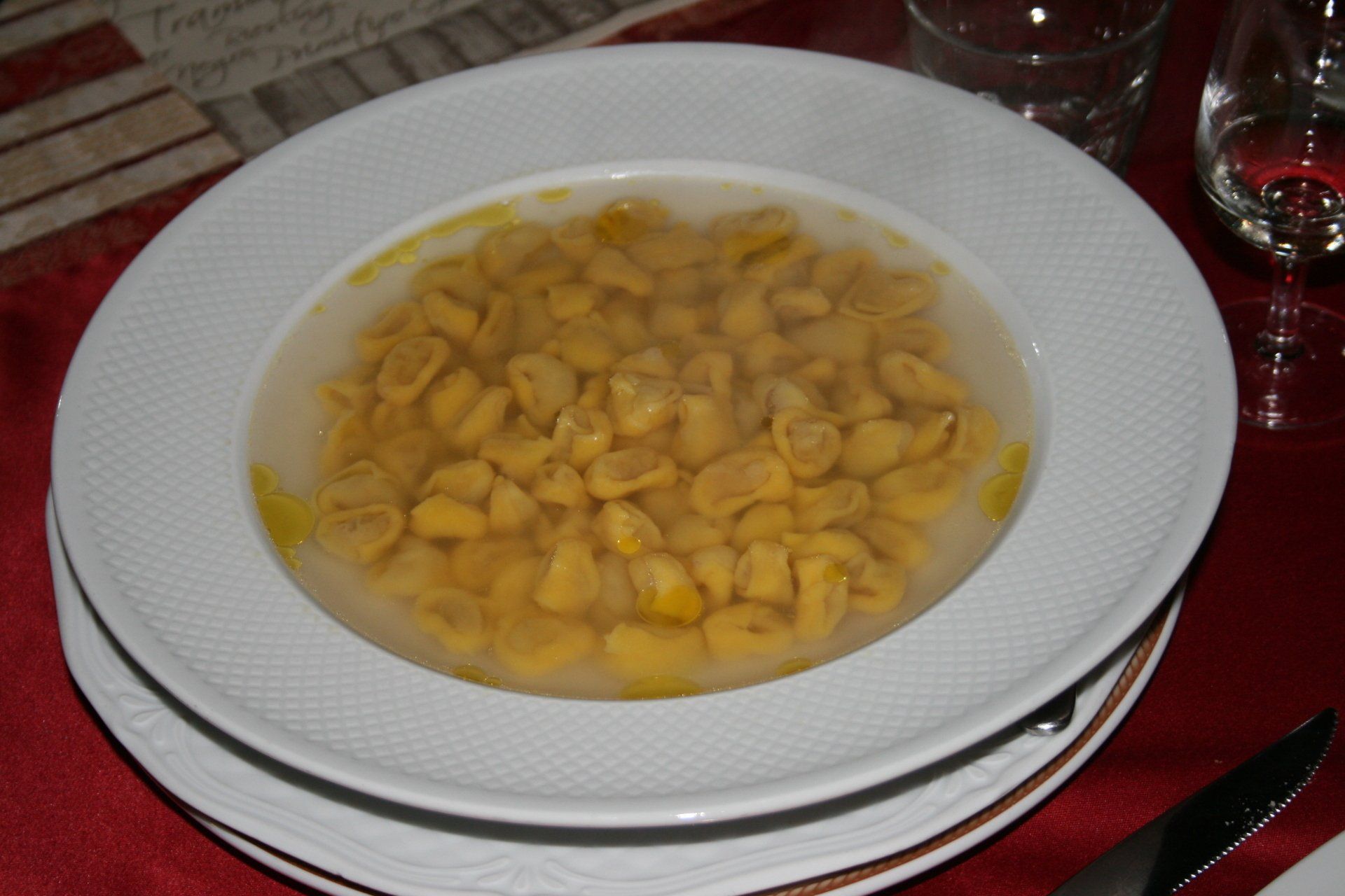 Ciotola di tortellini in brodo su un piatto bianco, posta su un tavolo con bicchieri.
