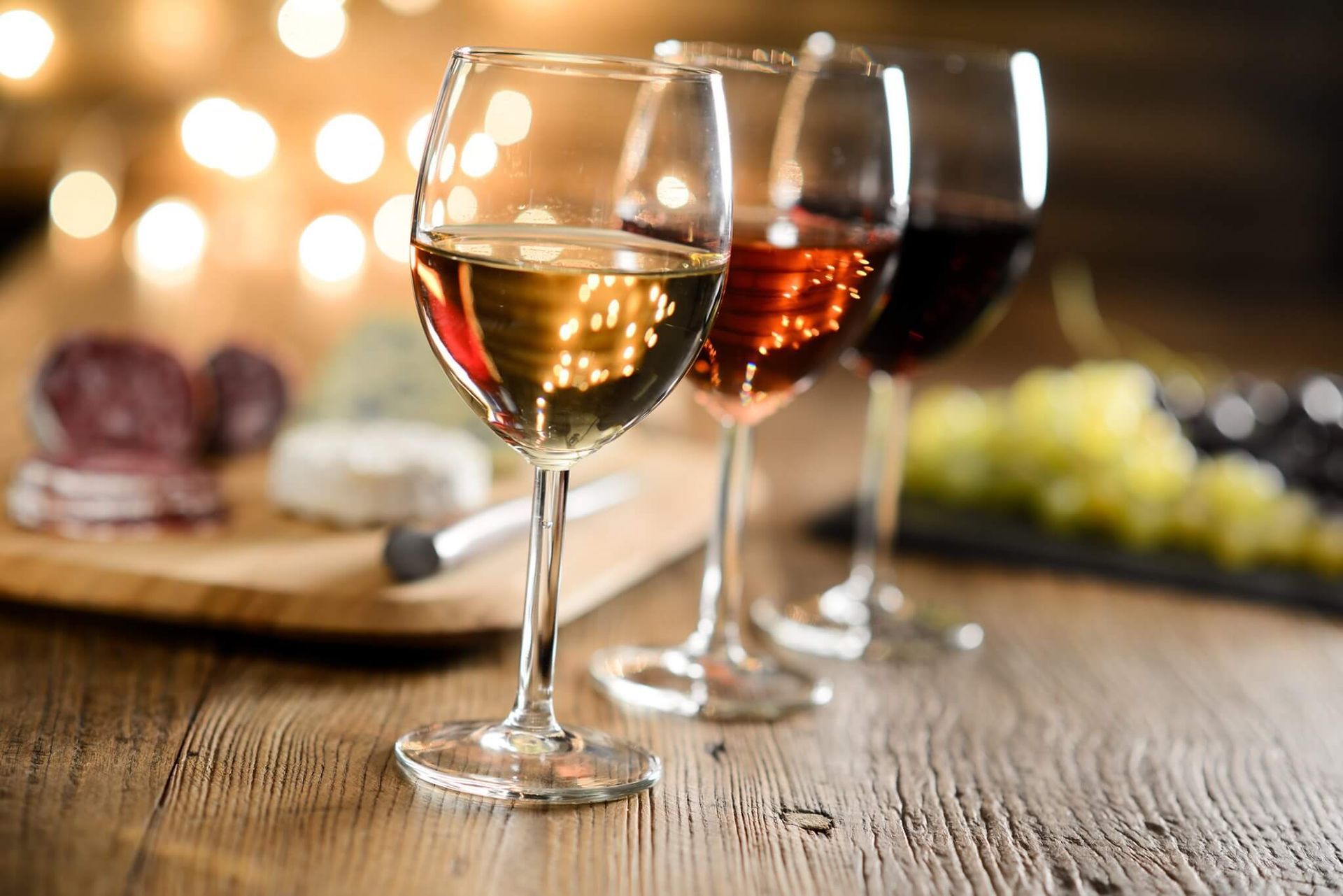 Tre bicchieri di vino bianco, rosato e rosso, accanto a un tagliere di formaggi e salumi, su un tavolo di legno.