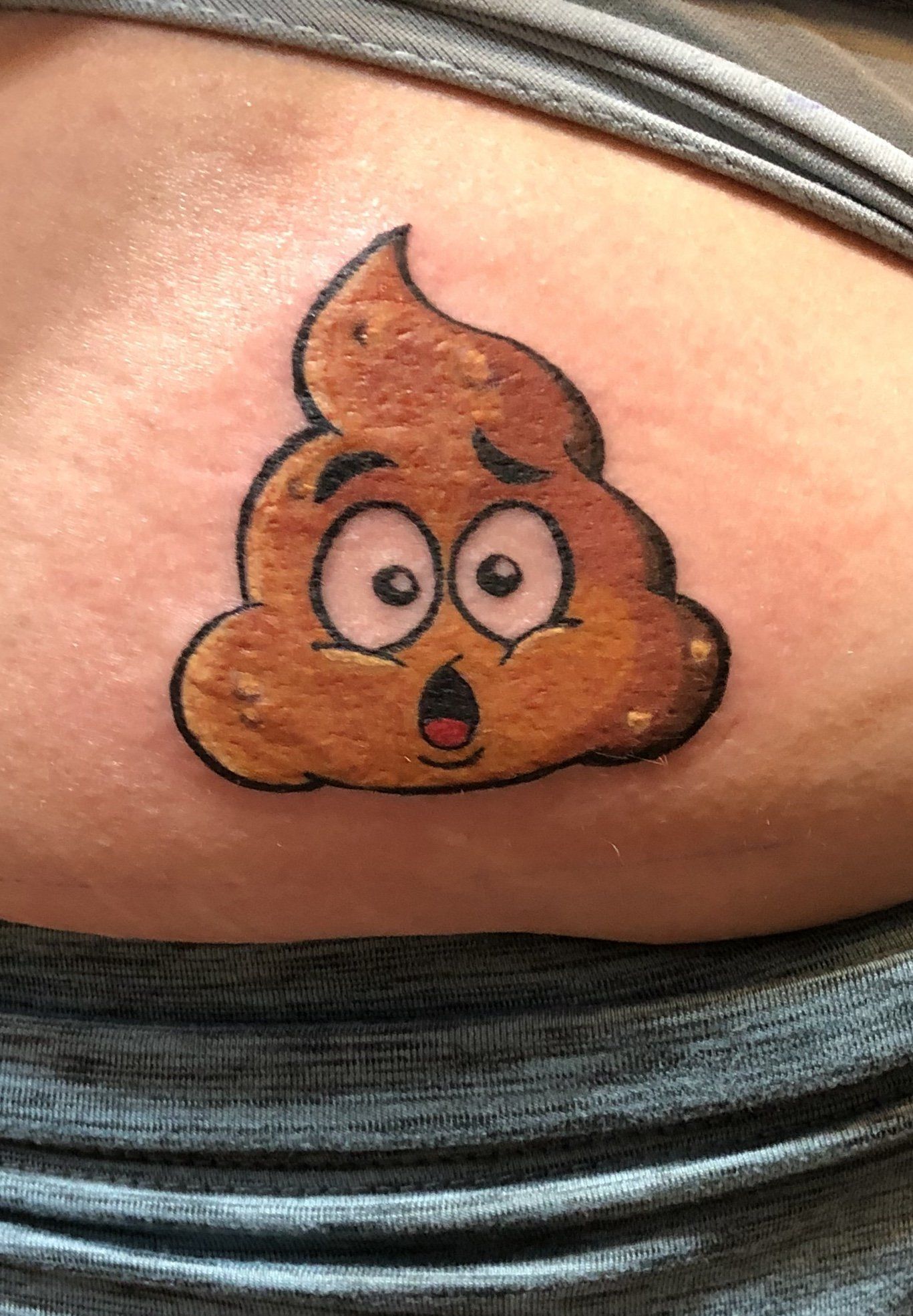 A cartoon poop emoji tattoo with surprised expression on someone's lower back, brown and tan, outlined in black.