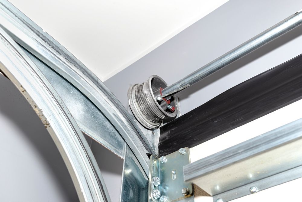 A metal garage door cable drum with a wound wire cable attached to a horizontal torsion bar above the door track.