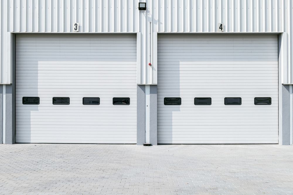 Two white industrial roll-up doors, labeled 3 and 4, each with a row of small windows, set in a light grey building.