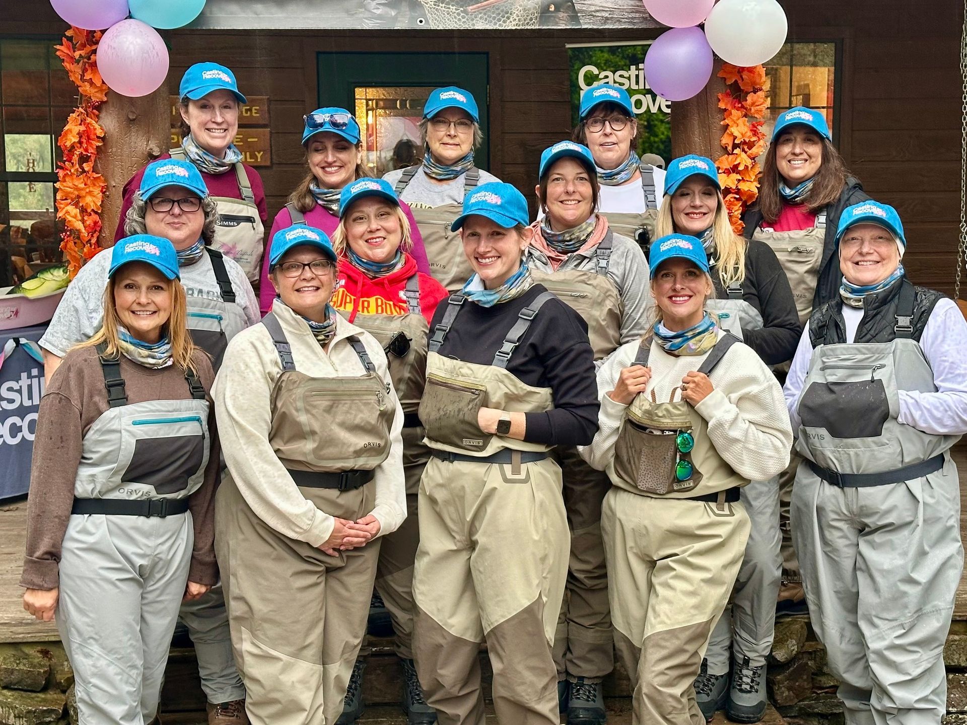 Group of people in blue hats and tan waders posing outside a storefront with balloons, smiling for a team photo
