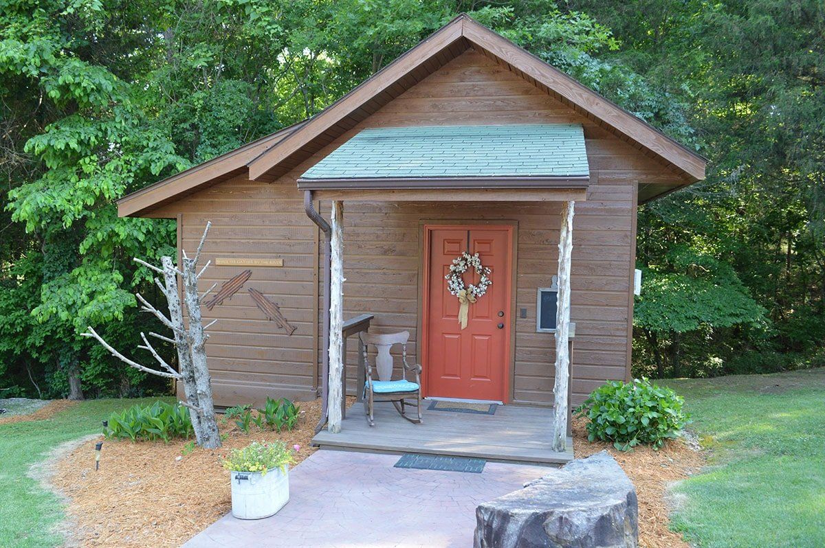 Small rustic wooden cabin with a red front door, porch, and surrounding greenery
