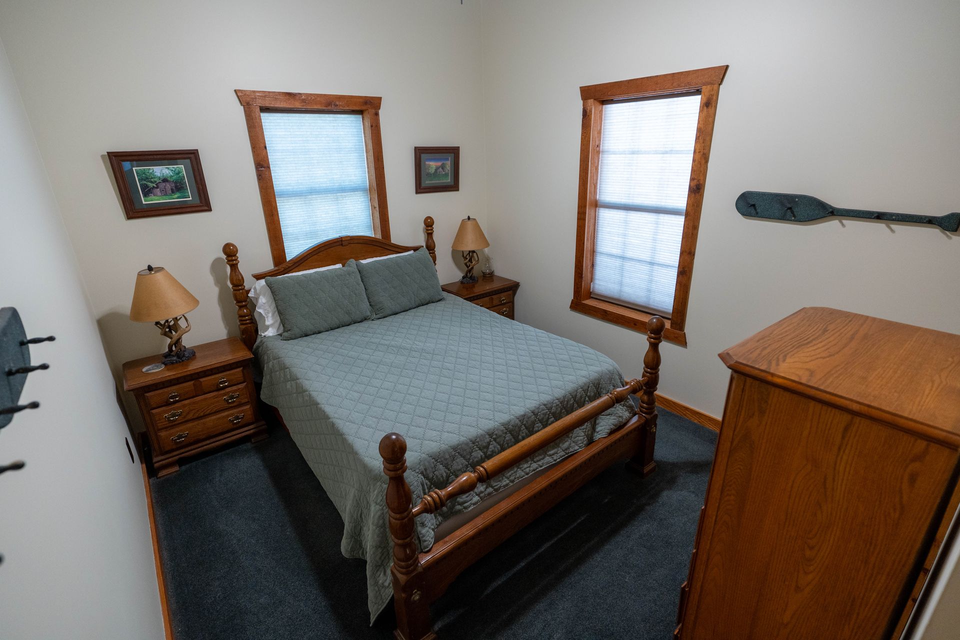Small bedroom with wooden bed, nightstand lamps, dresser, and two windows with blinds.