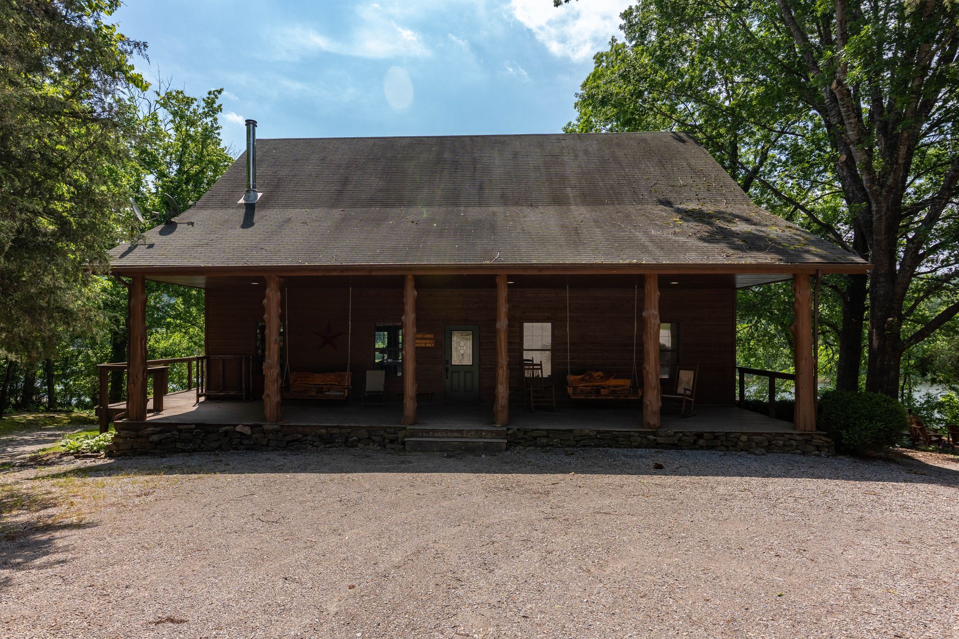 Brick pavilion with a wide porch and shingled roof, set in a gravel clearing among trees.