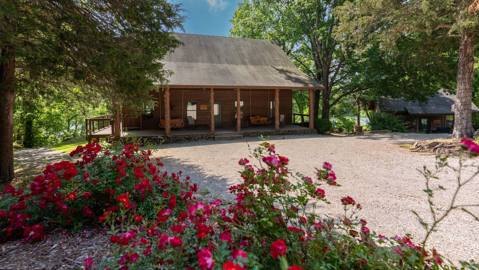Rustic cabin with a front porch amid trees, gravel driveway, and bright pink flowers in the foreground