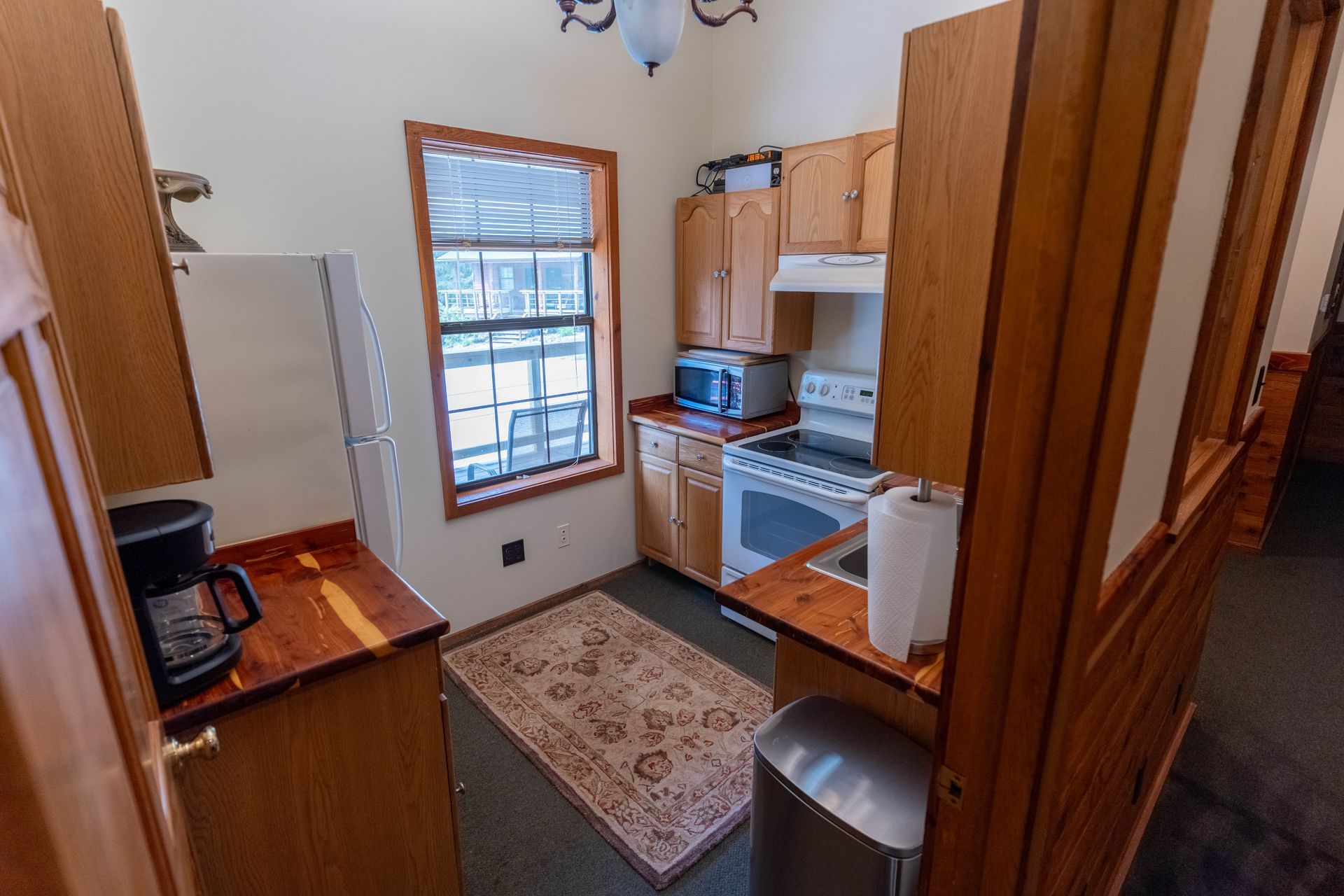 Small kitchen with wood cabinets, white appliances, rug, window, and coffee maker on the counter.