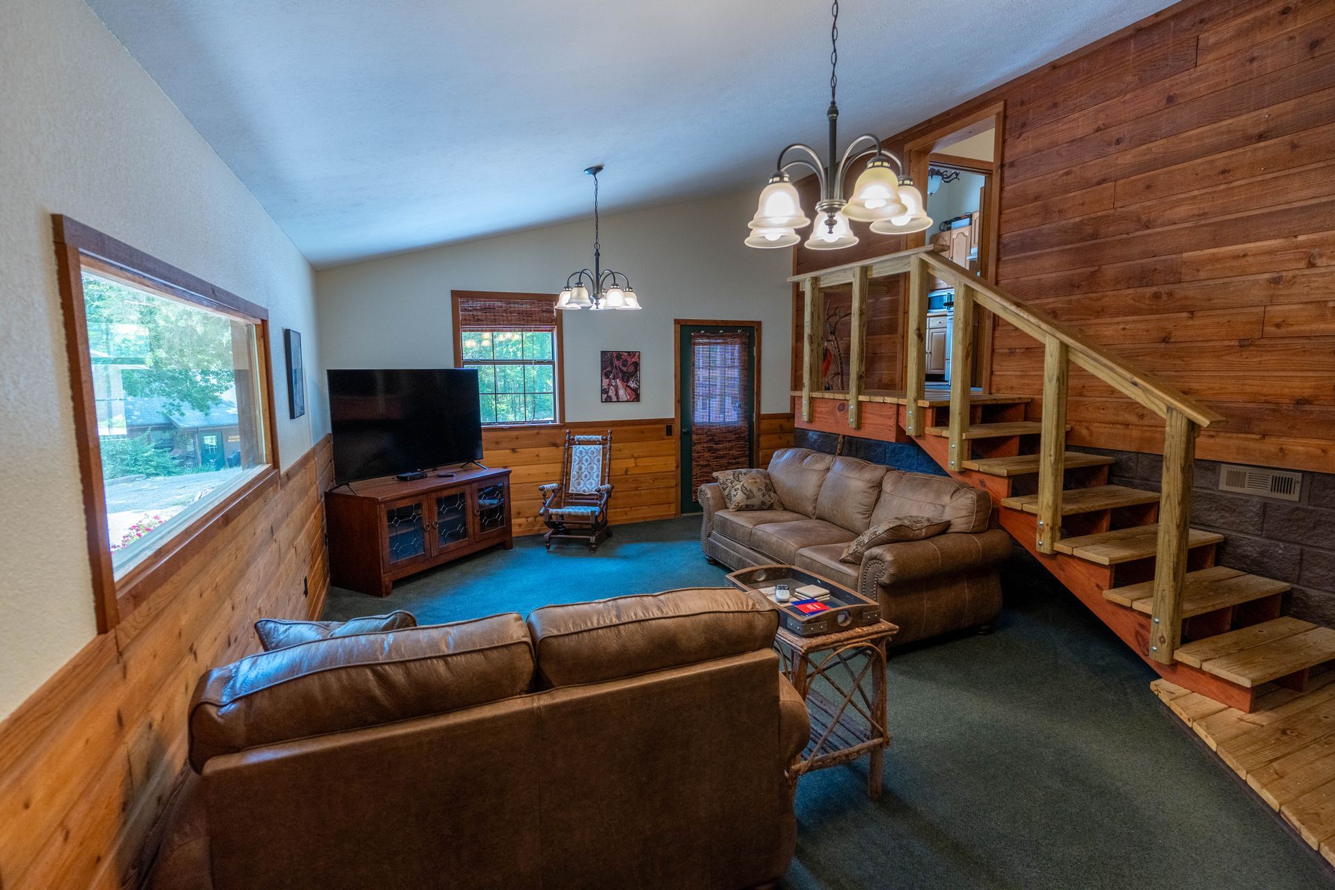 Cozy rustic living room with leather sofas, TV, wood-paneled walls, and staircase railing