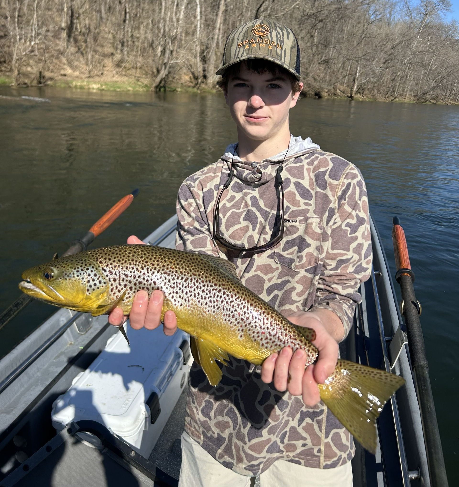 Person on a boat holding a large spotted fish on a riverbank, wearing camo gear.
