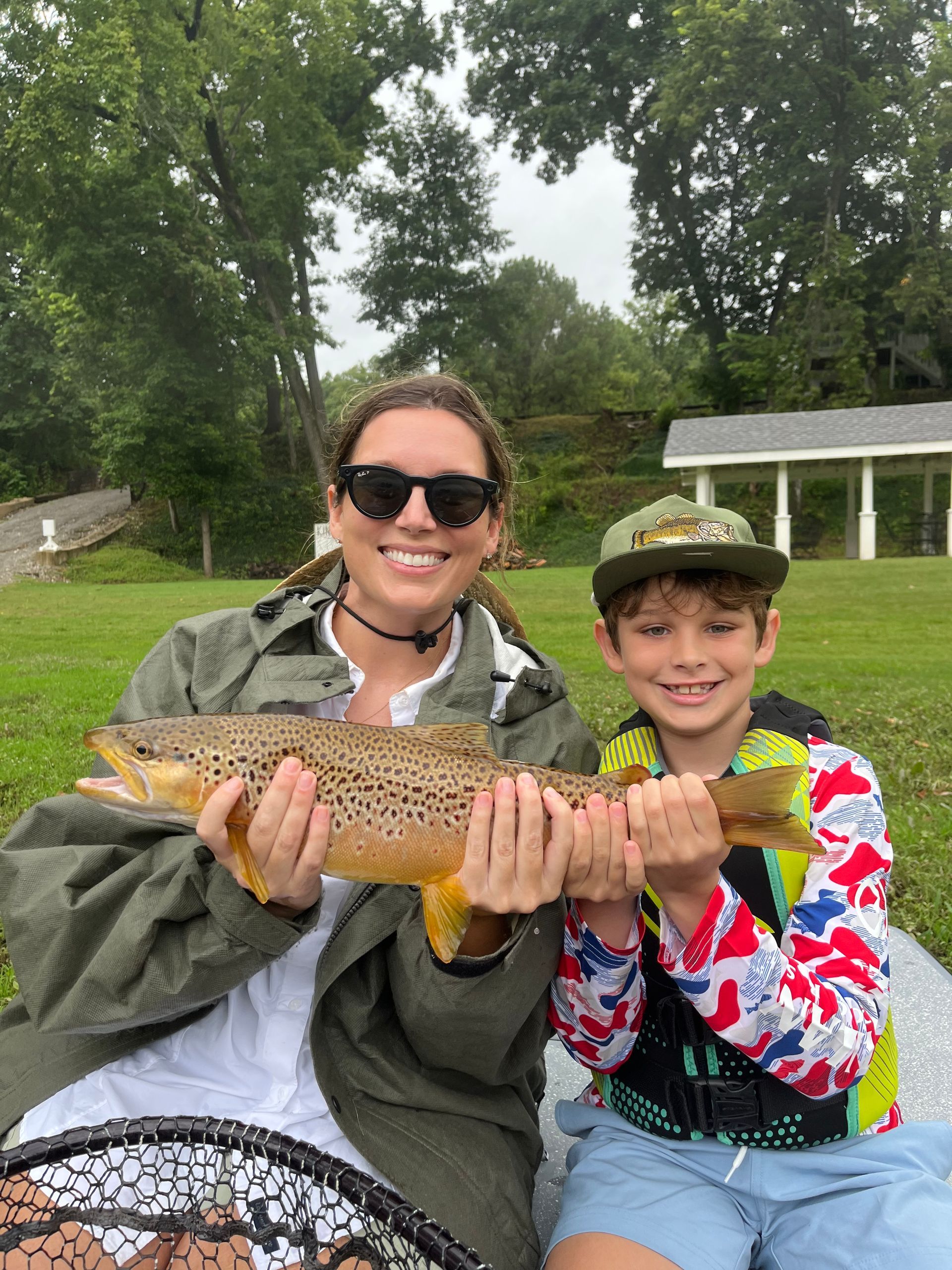 Two people smiling and holding a large fish outdoors by a lake