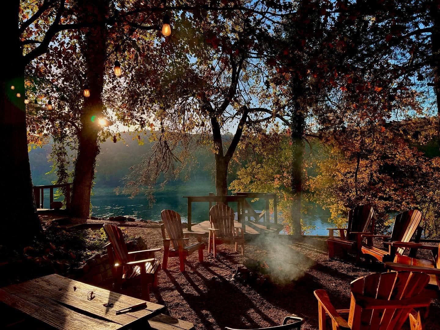 Sunlit outdoor patio with chairs and tables under trees, with lake or sea views in the background