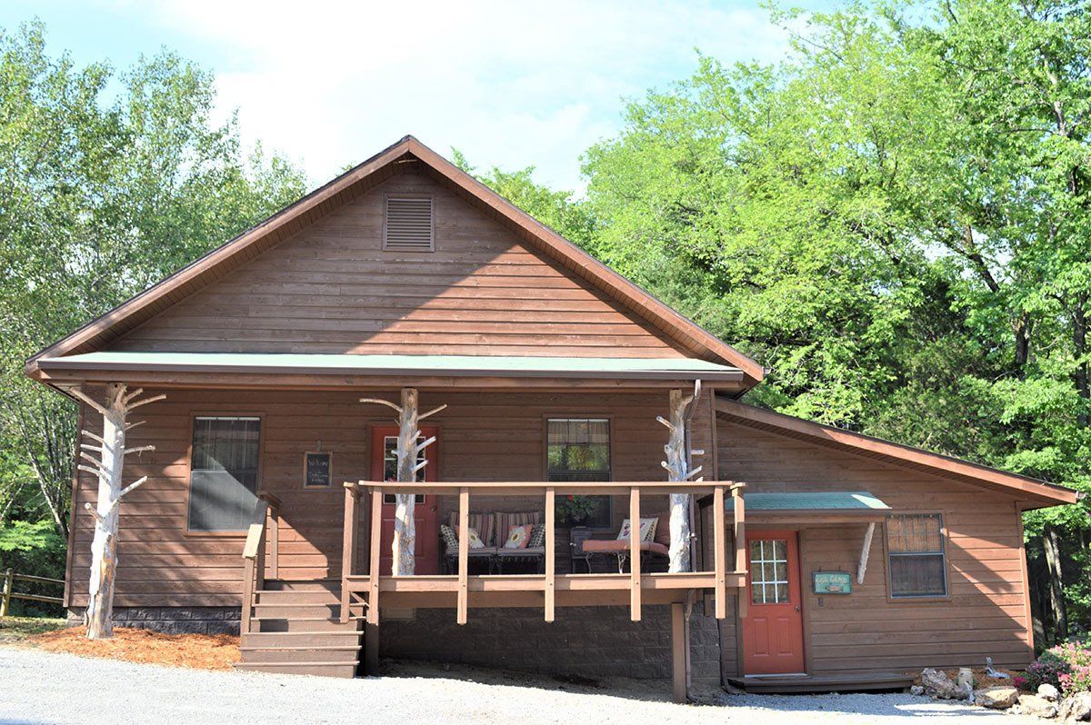Wooden cabin with a front porch and attached garage, surrounded by trees in bright daylight