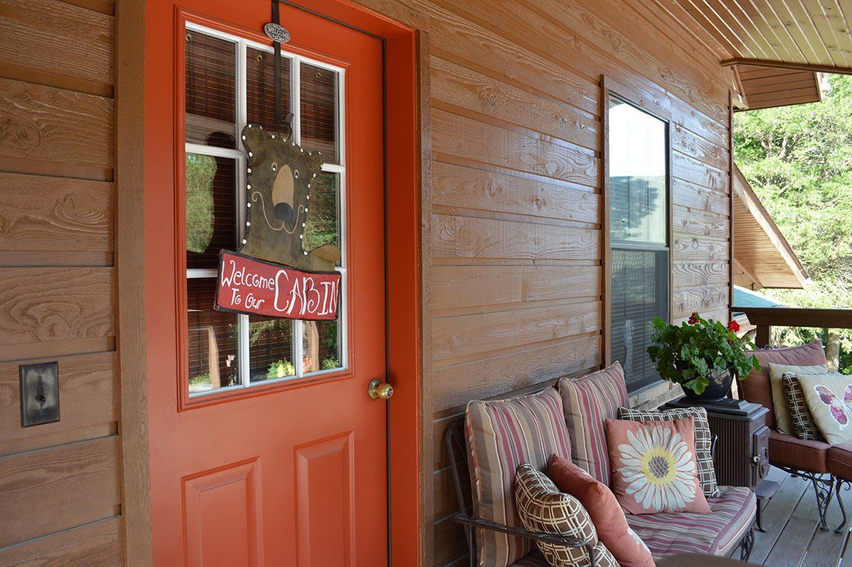 Orange porch door beside a cushioned bench and flowers on a wooden cabin porch