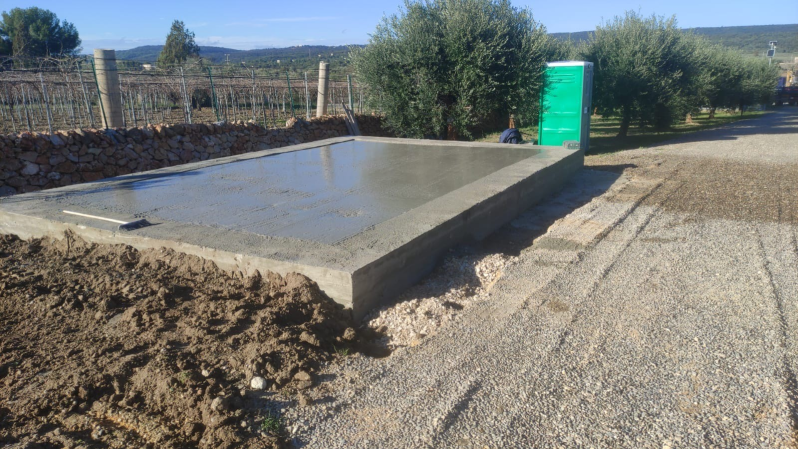 Base di fondazione in cemento accanto a un sentiero di ghiaia, con cumulo di terra, recinzione, cespugli e un bagno chimico verde sullo sfondo.