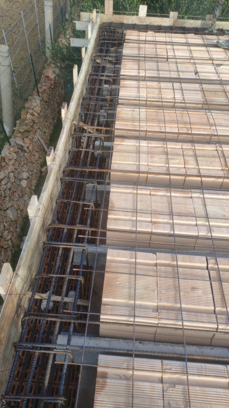 Rete di barre d'armatura e casseforme in legno in un cantiere edile accanto a una recinzione e un muro di mattoni.