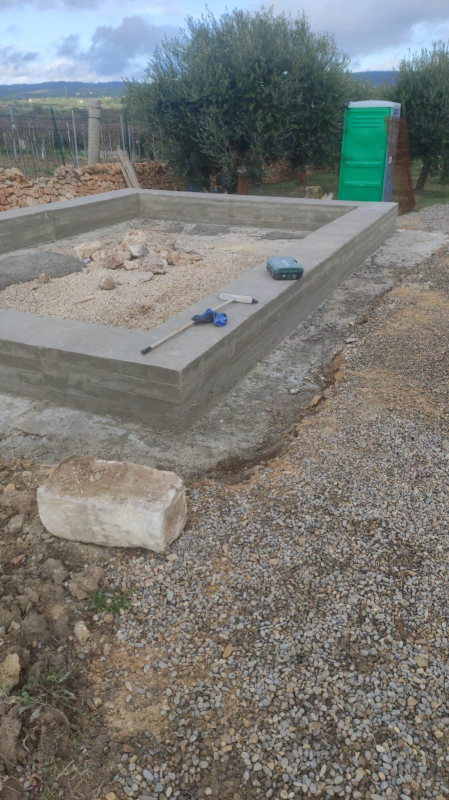 Soletta di fondazione in cemento in costruzione accanto a uno strato di ghiaia, con un bagno chimico verde sullo sfondo.