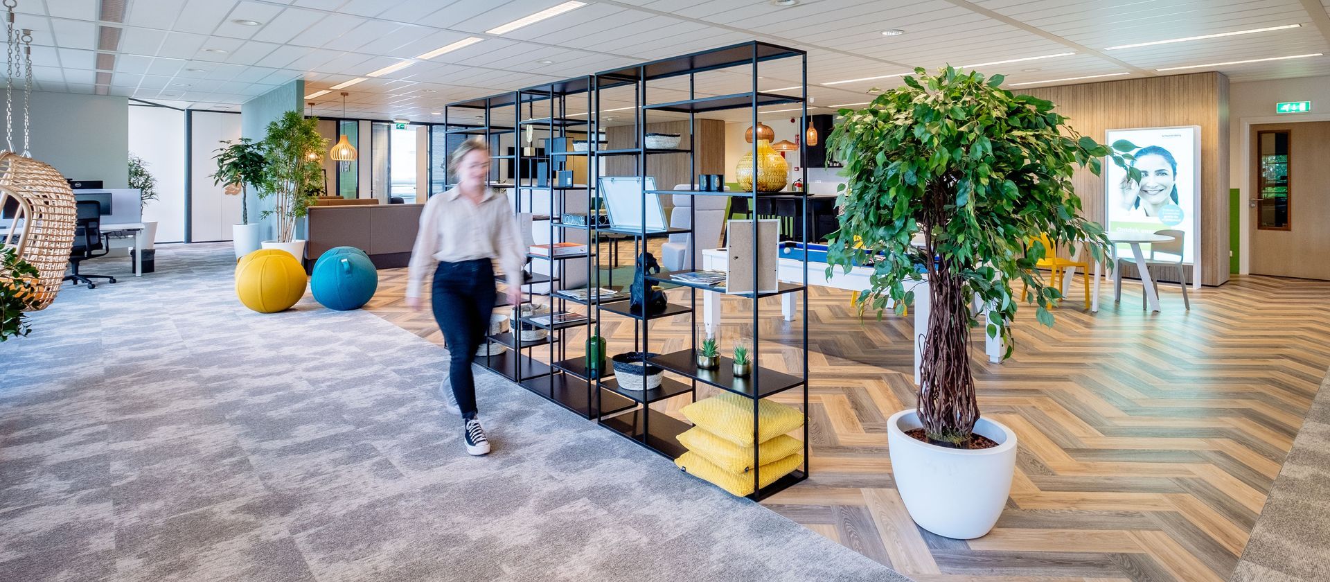 Modern office lobby with glass shelving, potted trees, colorful seating, and a person walking on patterned carpet