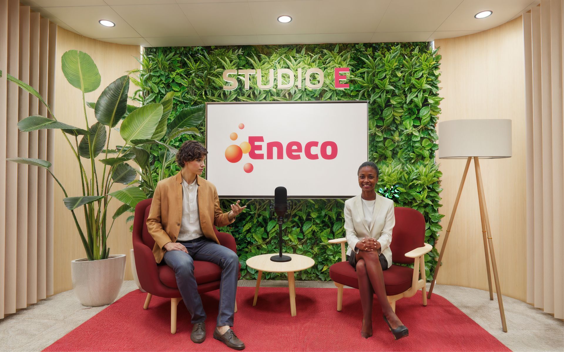 Twee mensen zitten op rode stoelen in een lichte studio met een Eneco-scherm en een groene plantenwand.