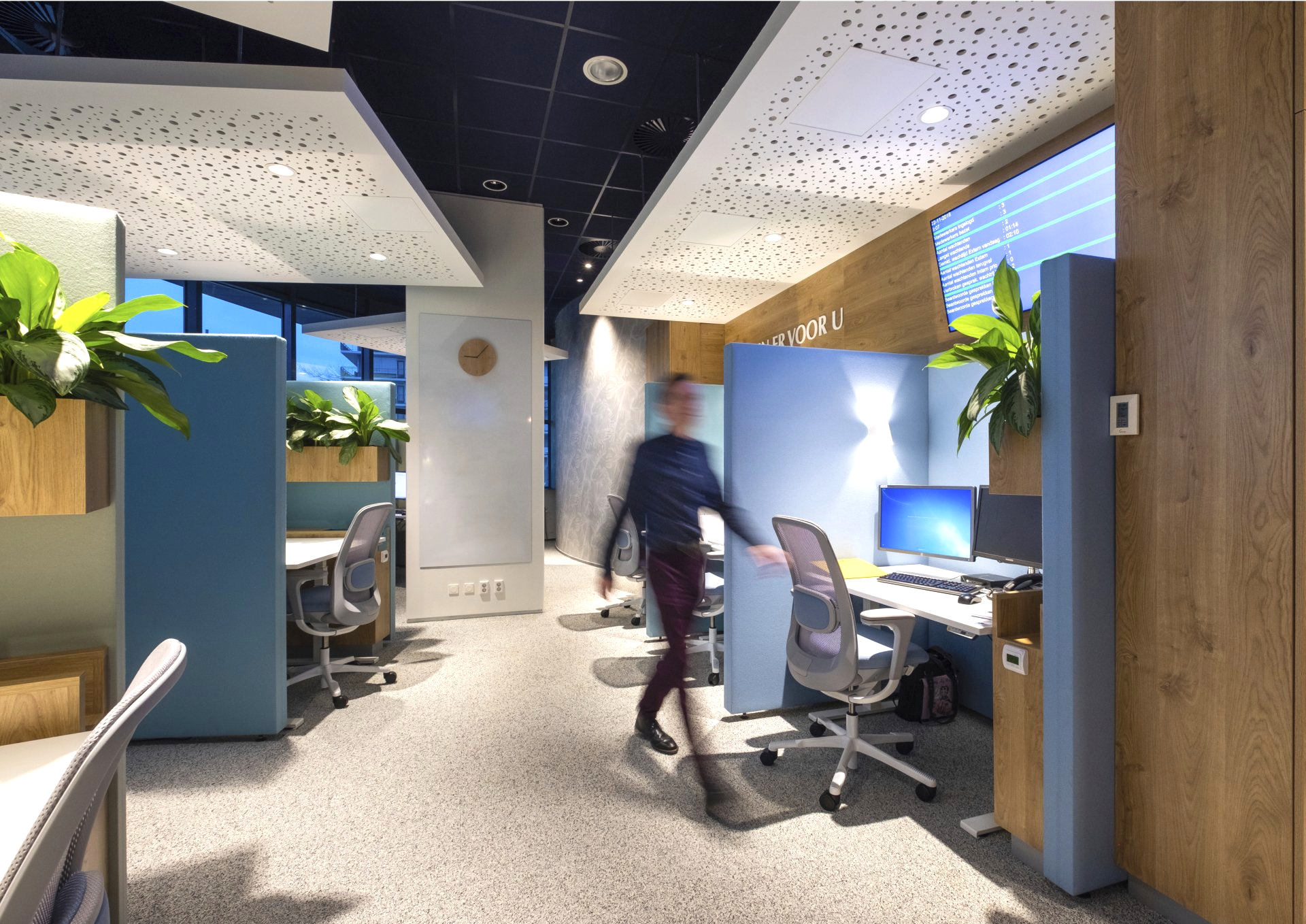 Moderne kantoorgang met bureaus, stoelen, planten en een persoon die langs verlichte werkplekken loopt.
