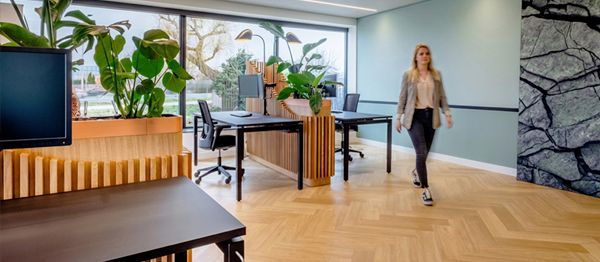 Moderne kantoorlounge met houten meubels, planten en een vrouw die op de achtergrond loopt.