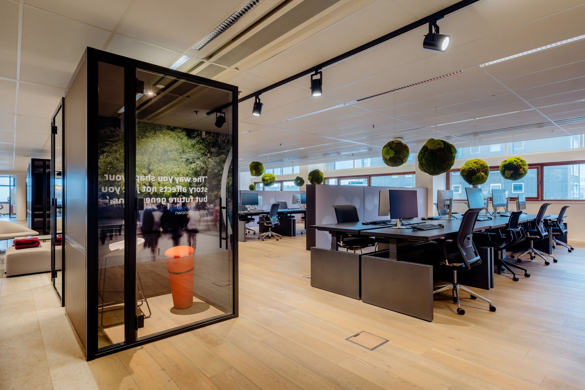 Modern open-plan office with desks, chairs, pendant lights, and a glass phone booth with plants inside