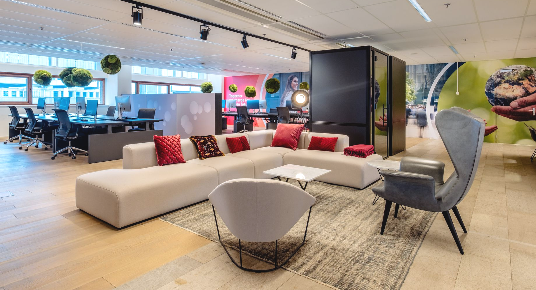 Moderne kantoorlounge met beige hoekbank, rode kussens, stoelen, vloerkleed en een open werkplek op de achtergrond.