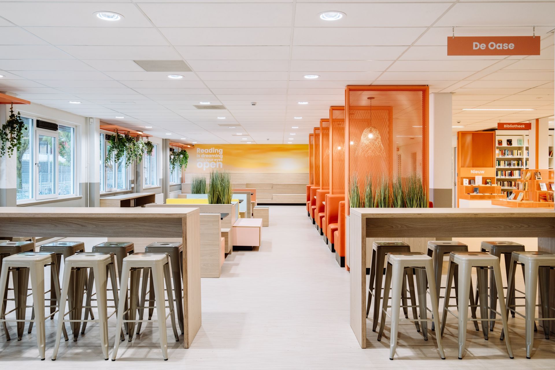 Een moderne school bibliotheeklounge met twee rijen barkrukken tegenover balies en een rij oranje, semi-privé zitjes. Vitaal leer- & leesplein
