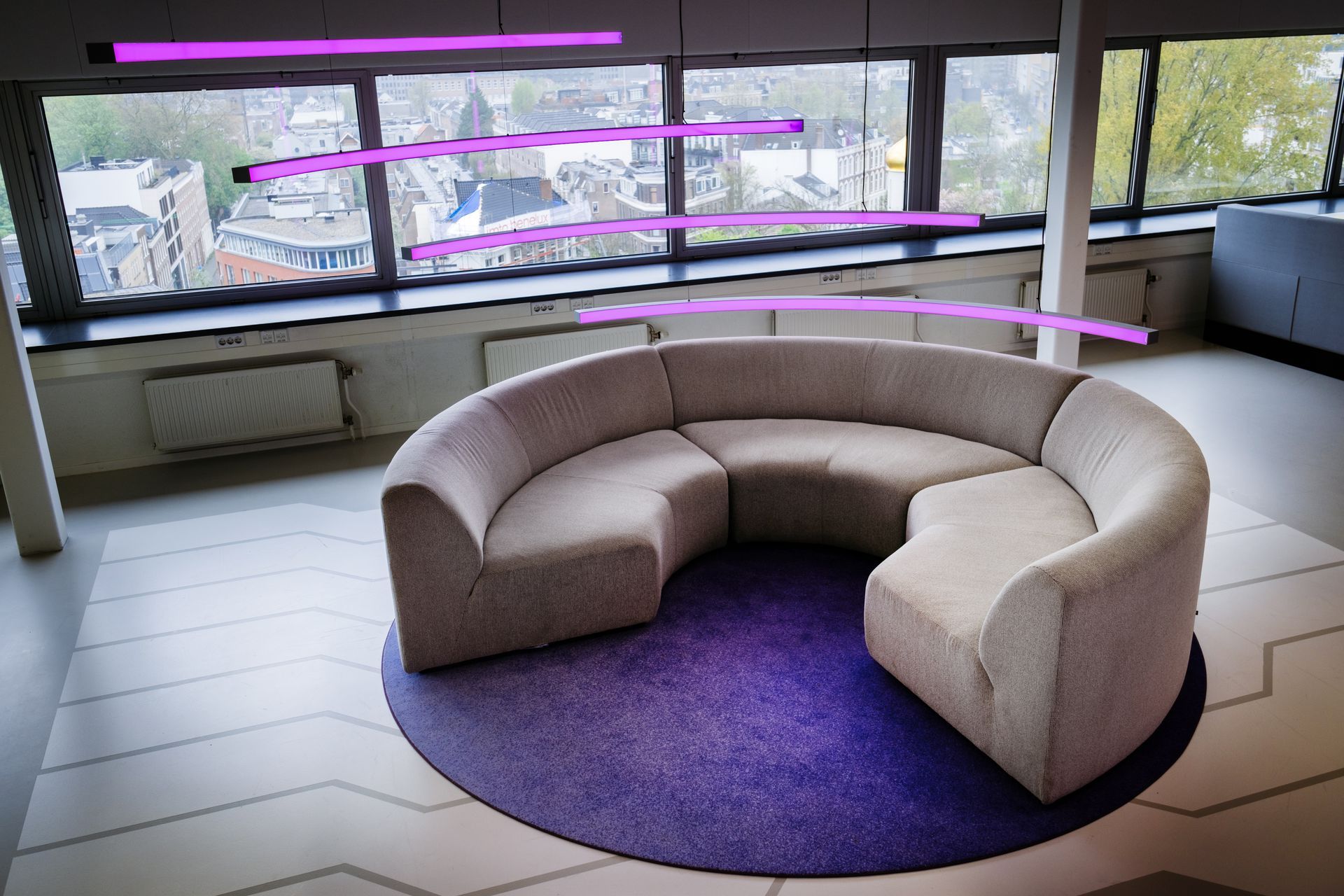 Een gebogen, beige modulaire bank staat op een rond paars vloerkleed in een kantoorruimte met grote ramen en paarse lichtelementen in het nieuwe kantoor van Shypple in Rotterdam, interieurontwerp voor KuóStudio