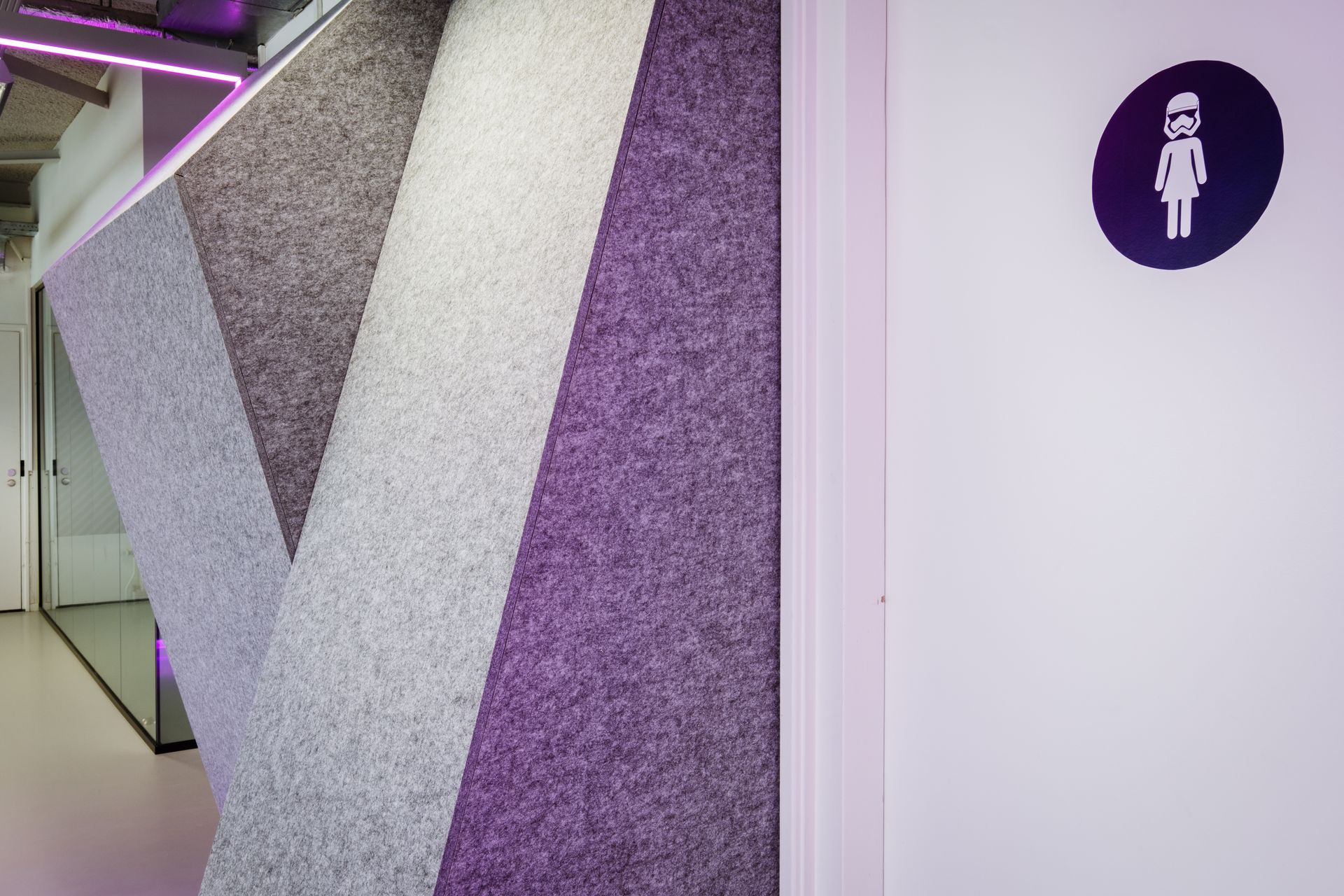 Een paars bord met een wit pictogram van een damestoilet, naast een muur met gestructureerde, hoekige akoestische panelen in grijs, in het nieuwe kantoor van Shypple in Rotterdam, interieurontwerp voor KuóStudio