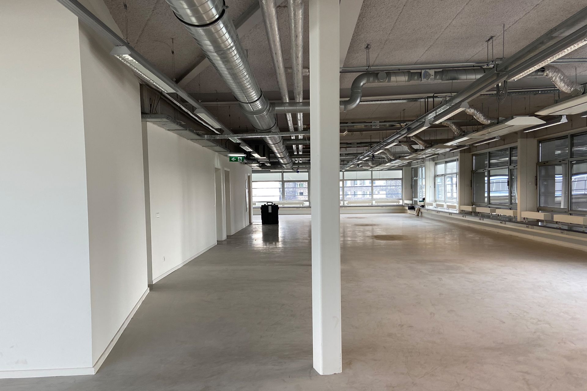 Voor verbouwing: Een lege, onafgewerkte kantoorruimte met zichtbare industriële leidingen, witte muren, grote ramen en een betonnen vloer in het kantoor van Shypple