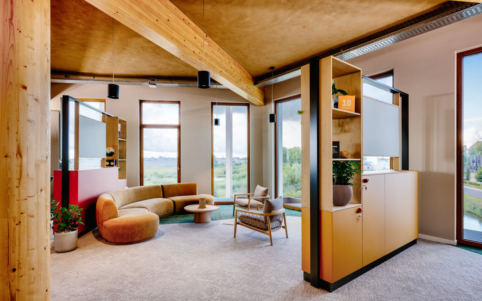 Moderne, open woonkamer met houten balkenplafond, grote ramen en beige zitmeubelen rond een stoel en een salontafel.