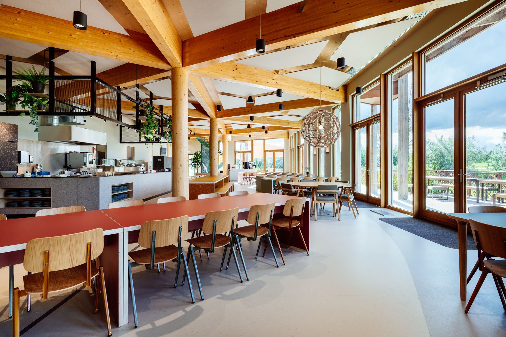 Een werkcafé met houten draagbalken, rijen stoelen aan tafels met gekleurde bladen en grote ramen die uitkijken op de buitenlucht  in het nieuwe kantoor van Bever, interieurontwerp door KuóStudio