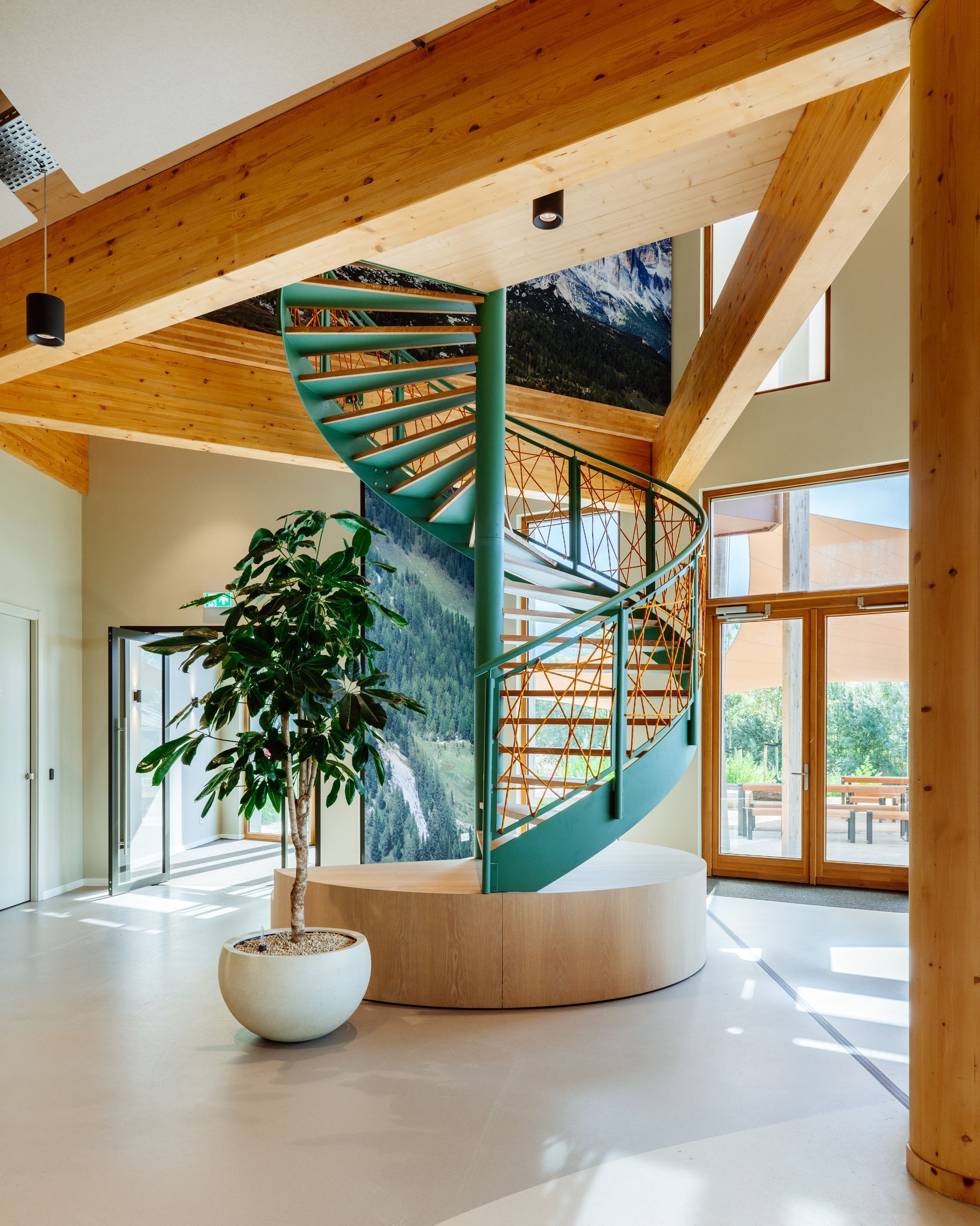 In het lichte, moderne werkcafé met houten balken en een ronde potplant staat een groene wenteltrap voorzien van klimtouw  in het nieuwe kantoor van Bever, interieurontwerp door KuóStudio.