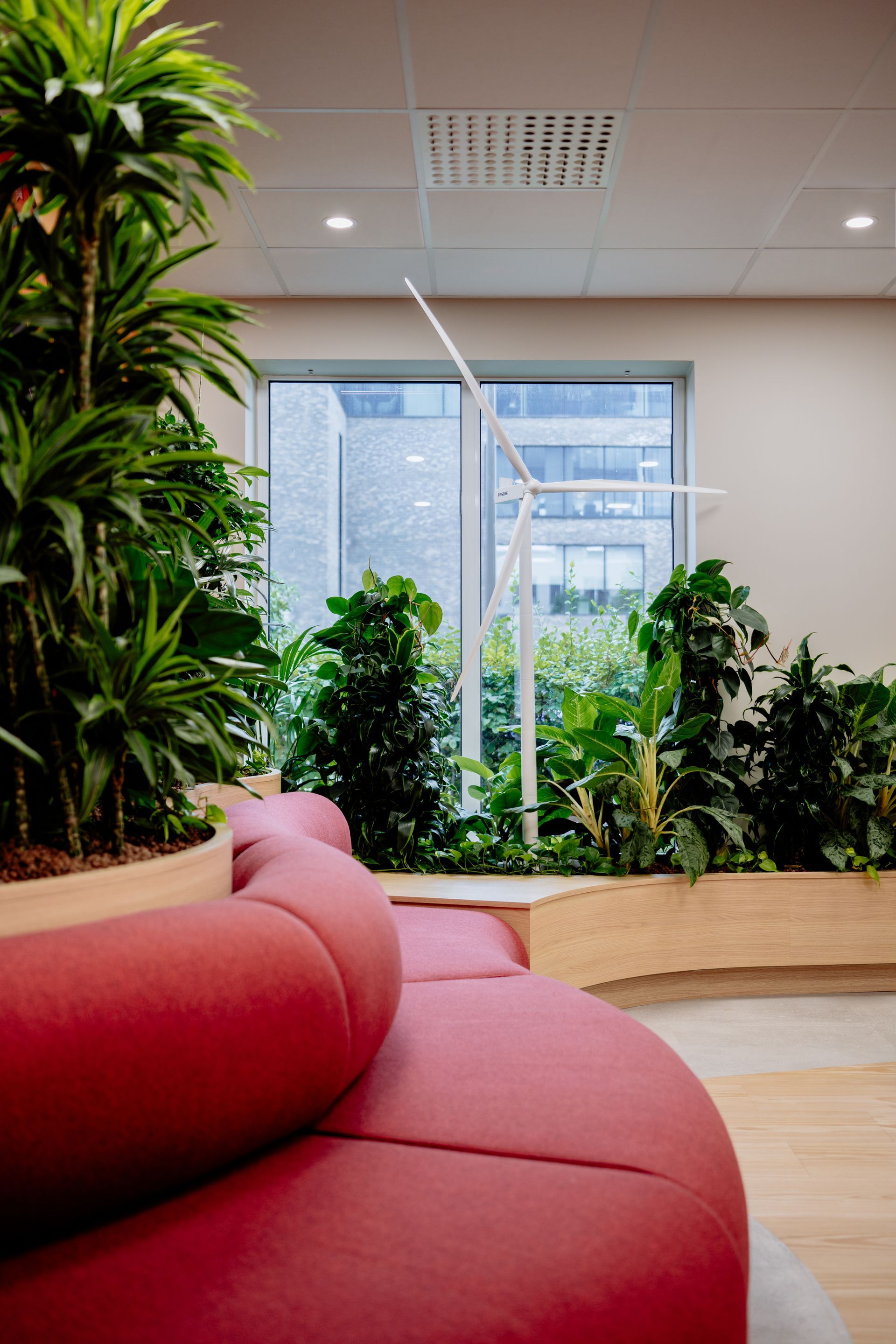 Moderne kantoorlounge met rode bank, hoge groene planten en grote ramen. Eneco Belgium
