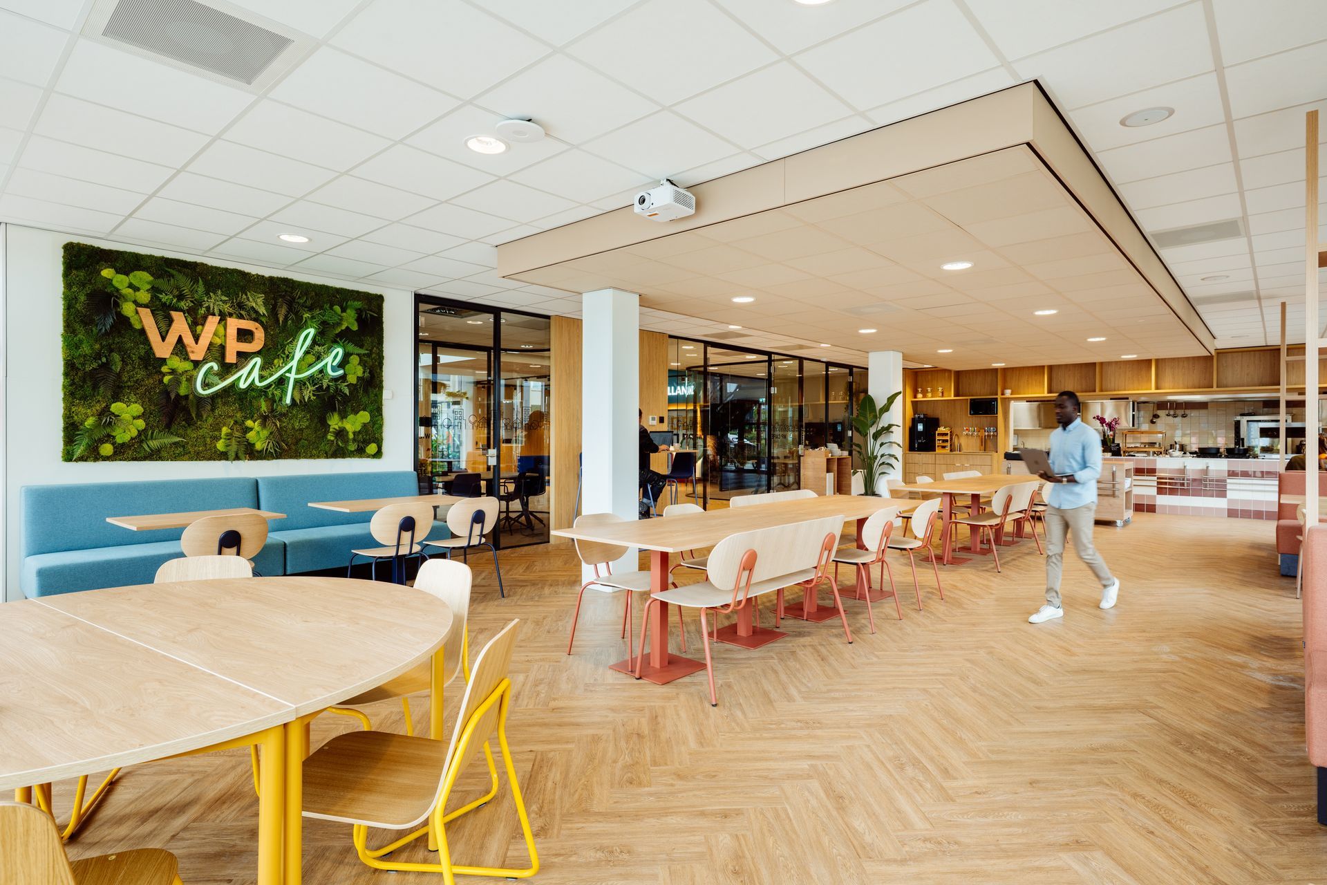 Modern café-interieur met houten tafels, blauwe stoelen, groene muurdecoratie en een voorbijlopende persoon.interieurontwerp door KuóStudio