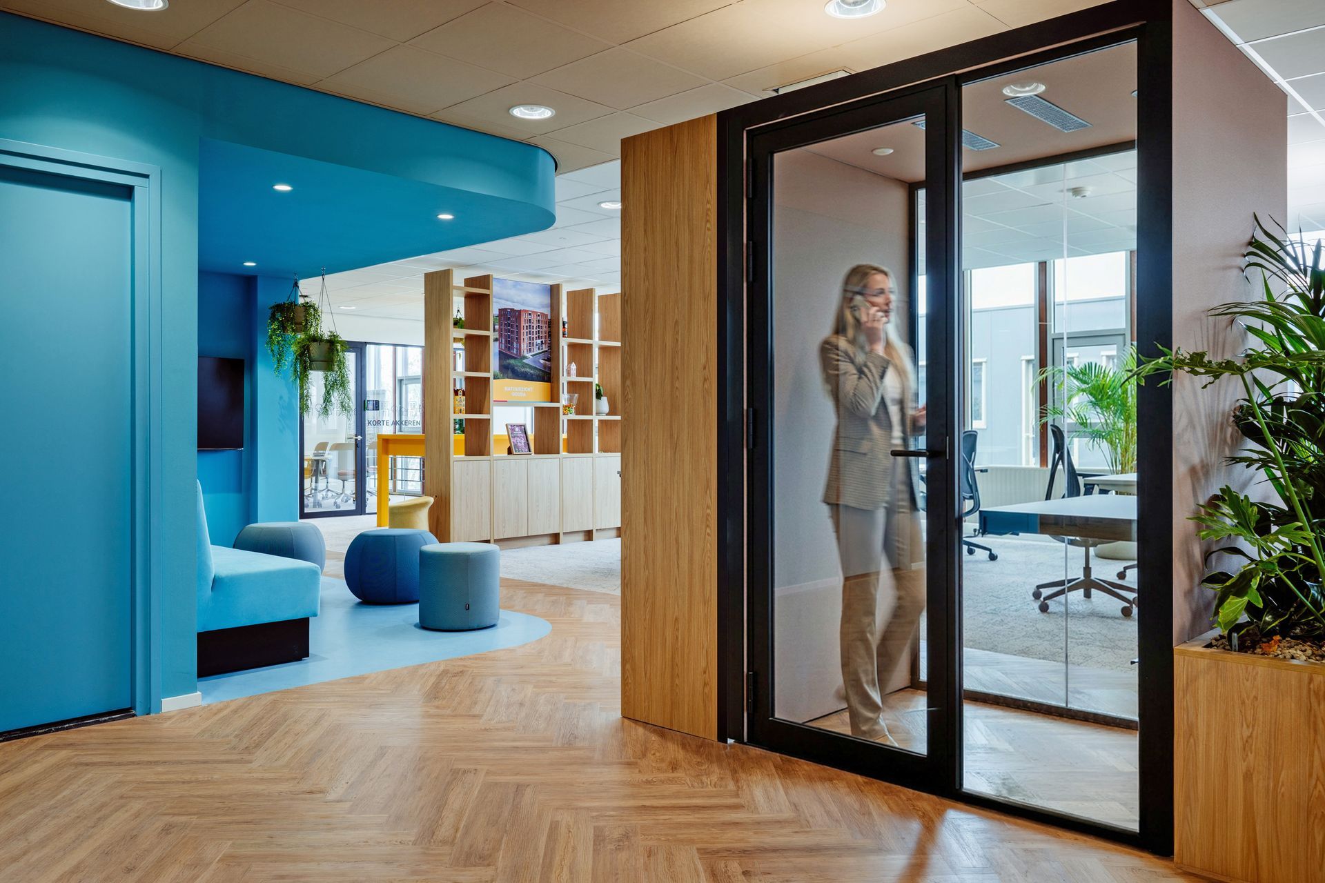 Een persoon staat in een glazen belcel van een kantoor in een moderne lobby met blauwe accenten en houten vloer. interieurontwerp door KuóStudio
